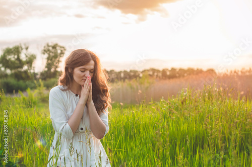 Girl closed her eyes, praying in a field during beautiful sunset. Hands folded in prayer concept for faith, spirituality and religion. Peace, hope, dreams concept