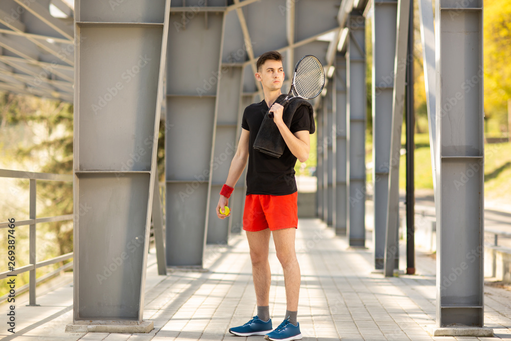 The man poses outdoors with a tennis racket and a ball. A towel is hanging on his shoulder. Sport concept
