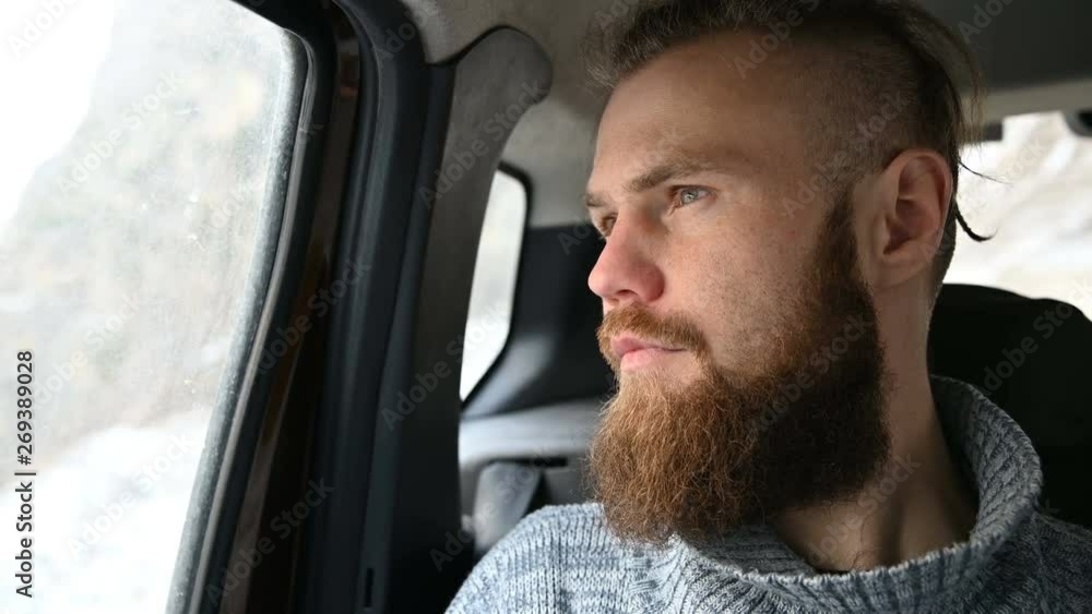 Close-up portrait of a bearded hipster male sitting in the car in the backseat and meditating while looking around Close-up portrait of a bearded hipster male sitting in the car in the backseat and meditating while looking around
