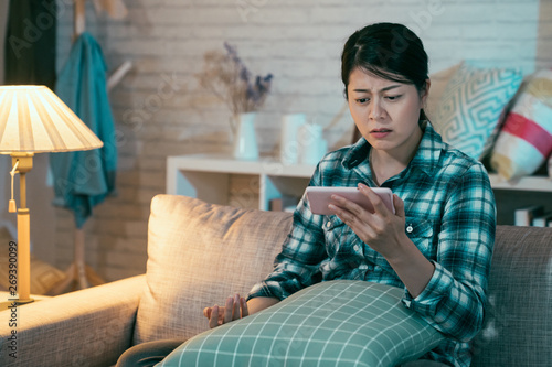 upset asian korean wife viewing online content in smart phone sitting on sofa in dark living room at home at night. sad girl binge watching love film romantic miserable movie on cellphone frowning. photo