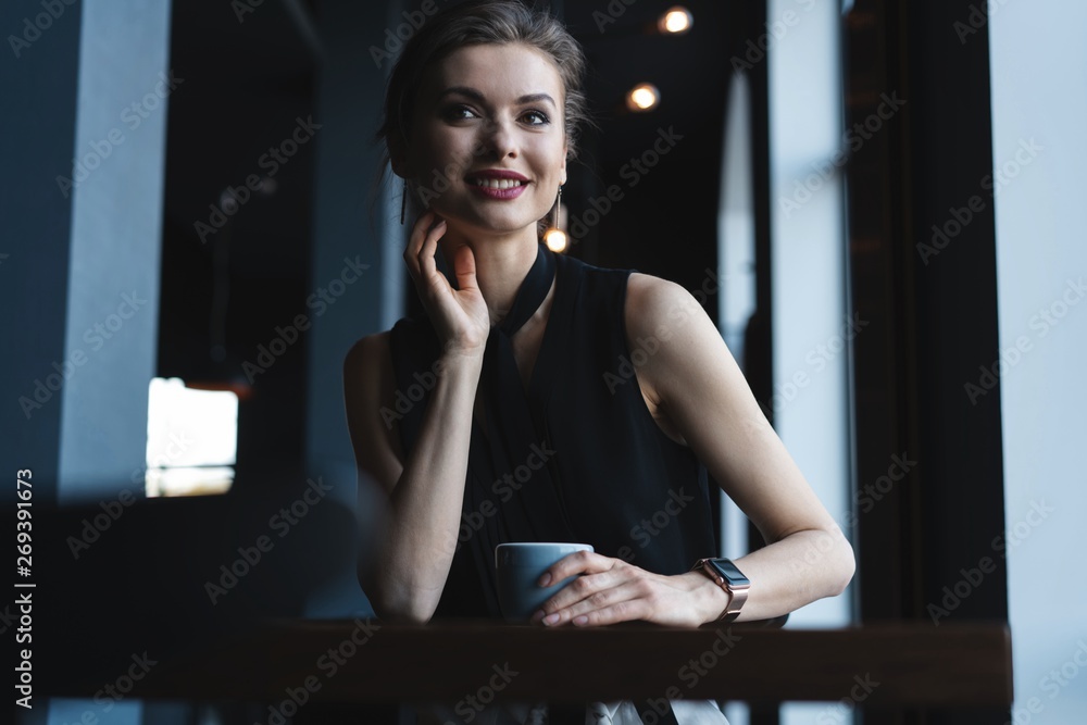 Portrait of gorgeous female drinking tea or coffee and looking with smile out of the coffee shop window while enjoying her leisure time, nice business woman lunch in modern cafe during her work break.