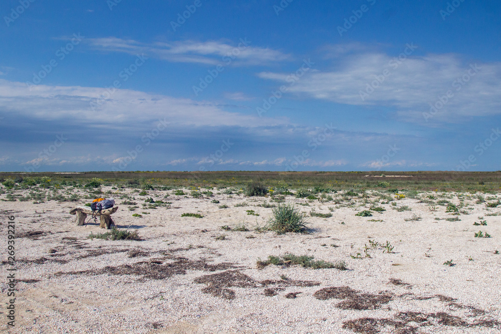 Wooden bench in the steppe. On the bench is a tourist backpack. Blue sky, land covered with small shells, steppe grass
