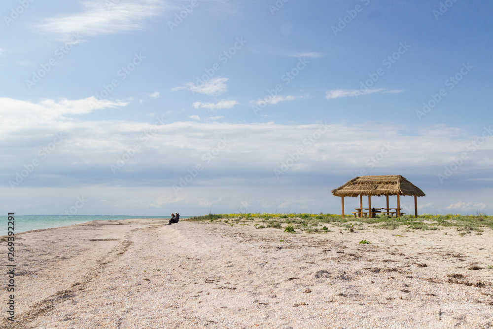 Sea shore. Gazebo with a roof of reeds. Someone is sitting on the beach. Steppe grass on the horizon.