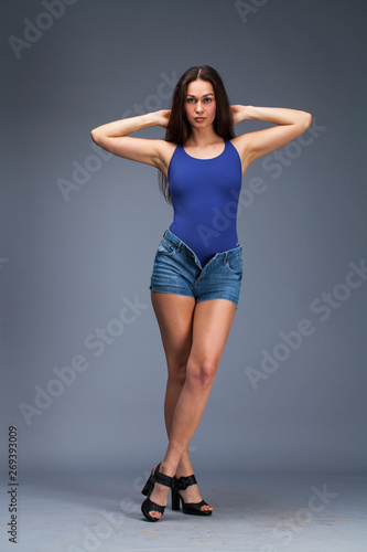 Brunette woman in blue bathing suit and jeans shorts posing in the studio on a gray background