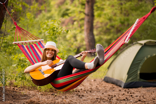 Woman in hat play guitar and relaxing in hammock hanging among pine trees in background. Camping rest concept
