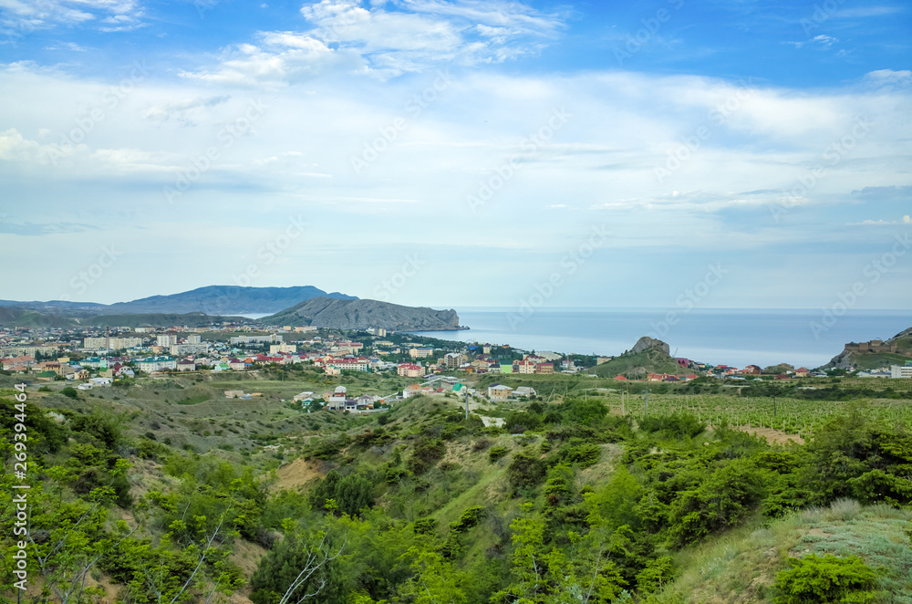 Sudak, Crimean Republic, Russia - May 30, 2017: View of the district of Sudak, residential buildings. Terrain in the mountains, vineyards, the sea on the horizon. View of Alchak mountain.