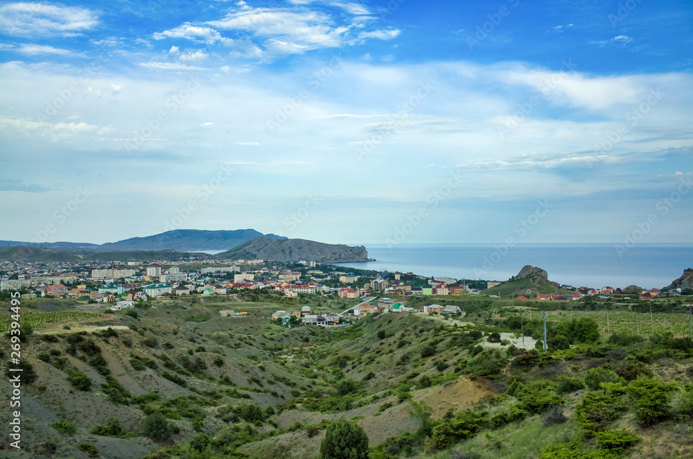 Sudak, Crimean Republic, Russia - May 30, 2017: View of the Sudak district, residential buildings, rolling hills, vineyards, sea on the horizon. View of Alchak mountain