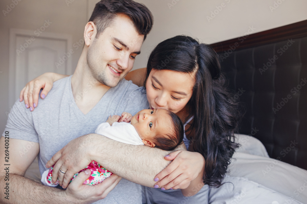 Portrait of beautiful smiling Caucasian father and Chinese Asian mother kissing mixed race newborn infant baby son daughter. Happy family in bedroom. Home lifestyle authentic natural moment. Portrait of beautiful smiling Caucasian father and Chinese Asian mother kissing mixed race newborn infant baby son daughter. Happy family in bedroom. Home lifestyle authentic natural moment.