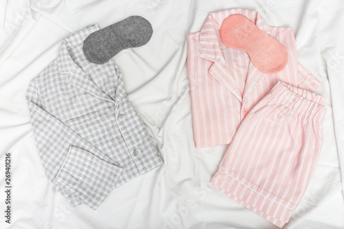 Two warm pajamas on bed, male and female, sleep masks. Gray and pink nightwear on white sheet. Top view. Flat lay. photo