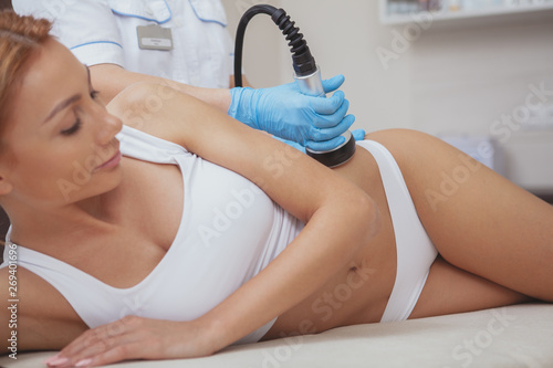 Cropped shot of a gorgeous woman with sexy body getting lifting body treatment by cosmetologist. Beautician using hardware cosmetology machine on a female client, doing ultrasound cavitation photo