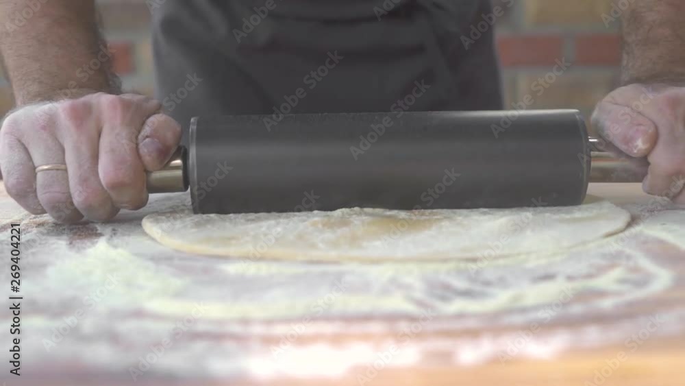 Cook sprinkling flour and rolling thin dough with roller pin at kitchen table. Husband cooking pie dough on home kitchen. Preparing italian pizza in restaurant. Cooking handmade food concept. Cook sprinkling flour and rolling thin dough with roller pin at kitchen table. Husband cooking pie dough on home kitchen. Preparing italian pizza in restaurant. Cooking handmade food concept.