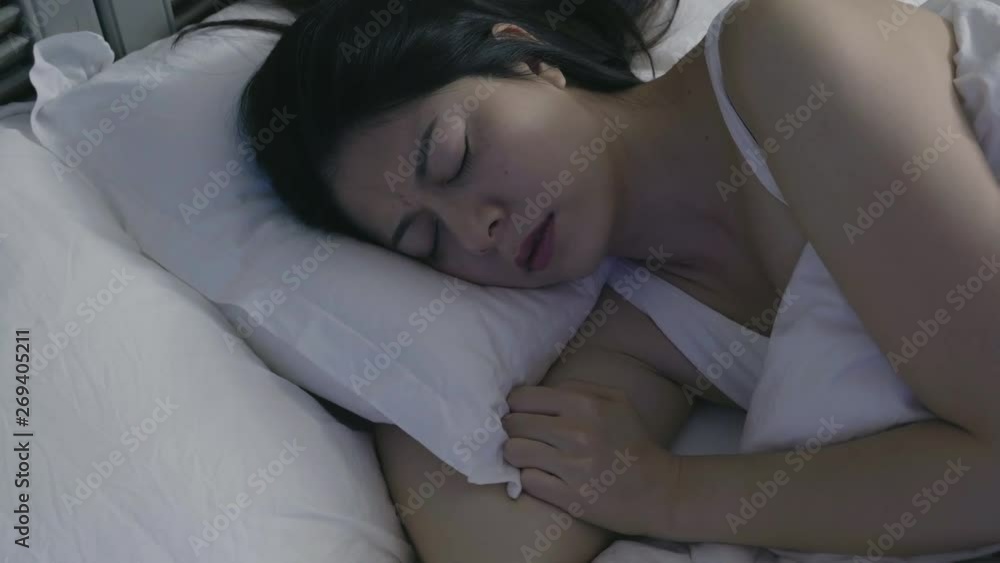 slow motion of scared asian lady sit up with frowning face hand touching hair forehead. Restless dreams of sleeping woman interrupted by waking up for nightmares from white bed in bedroom at night. slow motion of scared asian lady sit up with frowning face hand touching hair forehead. Restless dreams of sleeping woman interrupted by waking up for nightmares from white bed in bedroom at night.
