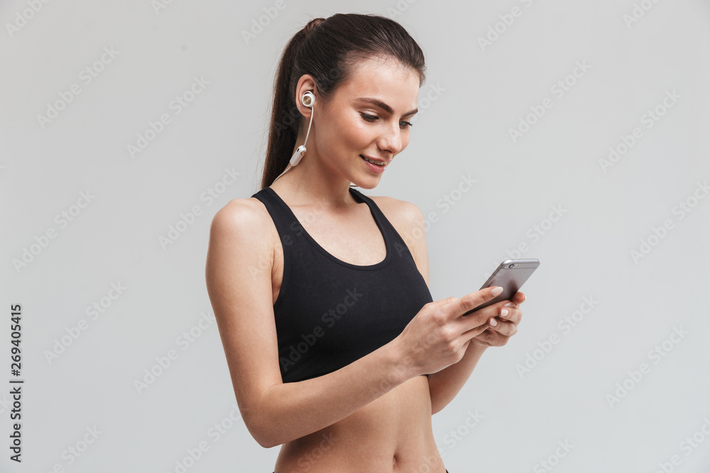 Beautiful young sport fitness woman runner using mobile phone listening music isolated over grey wall background. Beautiful young sport fitness woman runner using mobile phone listening music isolated over grey wall background.