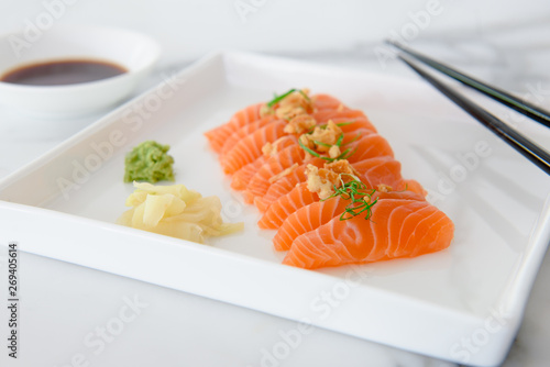 Sliced salmon sashimi, a popular Japanese food, on white plate with wasabi, pickled ginger and soy sauce on the side shown with chopsticks and white background