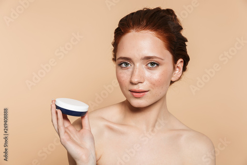 Amazing young redhead woman posing isolated over beige wall background take care of her skin with cream.