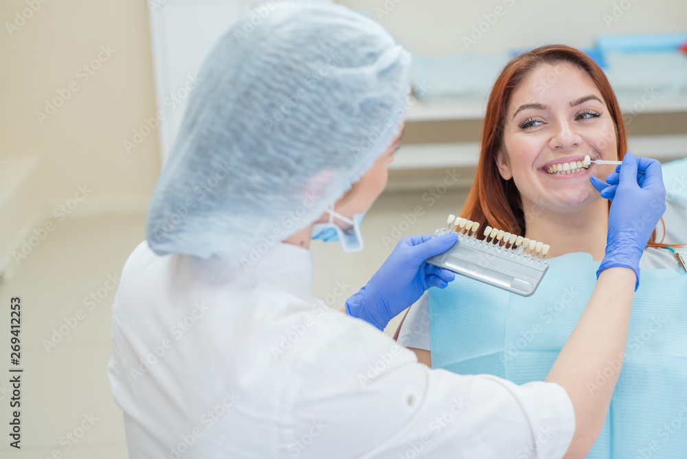 Naklejka premium Check and select the color of the teeth in the dentist chair. Dentist makes the process of treating a beautiful young red-haired girl. Selection of a tooth implant.