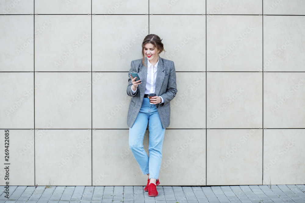 Adult woman stands near the wall with coffee. Communicate on the Internet. Concept of lifestyle, urban, business, study Adult woman stands near the wall with coffee. Communicate on the Internet. Concept of lifestyle, urban, business, study
