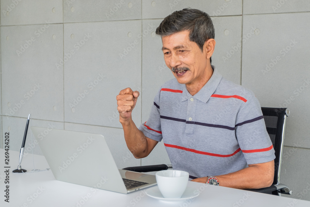Asian old man. Rejoice, raise fist right hand. Sitting on the chair look at to laptop computer screen, pen and white coffee mug on the desk in the office. Asian old man. Rejoice, raise fist right hand. Sitting on the chair look at to laptop computer screen, pen and white coffee mug on the desk in the office.