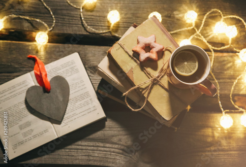 Holidays mood photo. Christmas lights and hot tea mug. Book for cosy evening. Sweet gingerbread and wooden heart on tray. Perfect winter flat lay with candle. Hygge concept.