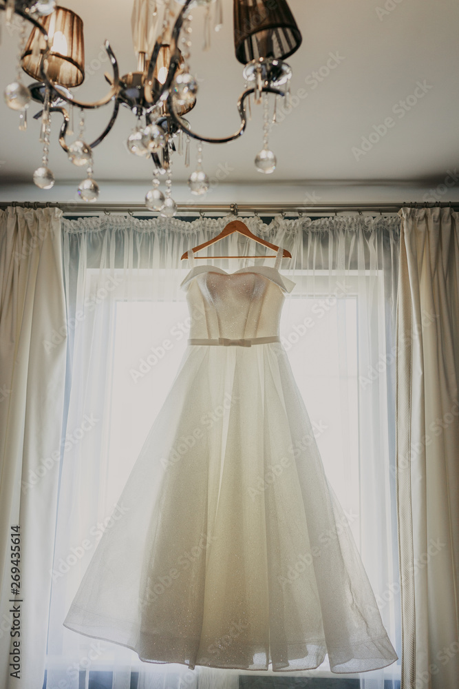 Wedding dress. White wedding dress with a full skirt on a hanger in the room of the bride with white curtains. Wedding attributes. No people. Wedding dress. White wedding dress with a full skirt on a hanger in the room of the bride with white curtains. Wedding attributes. No people.