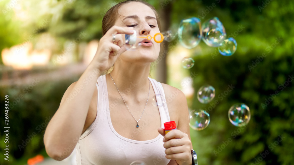 Portrait of happy smiling young woman blowing colorful soap bubbles at summer park