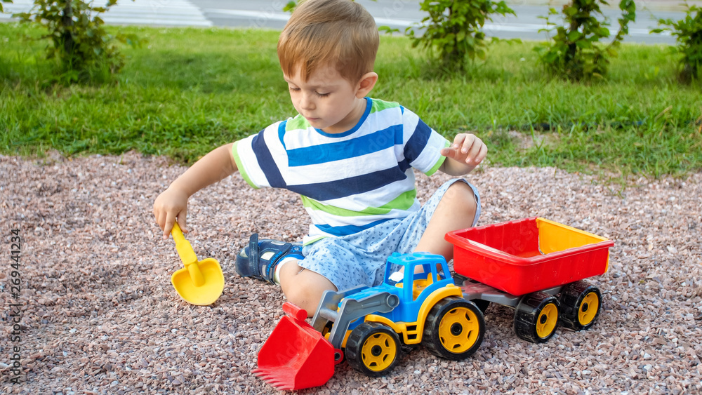 Portrait of cute 3 years old toddler boy sitting on the playground at park and playing with colorful plastic toy truck. Child having fun and playing outdoors with toys Portrait of cute 3 years old toddler boy sitting on the playground at park and playing with colorful plastic toy truck. Child having fun and playing outdoors with toys