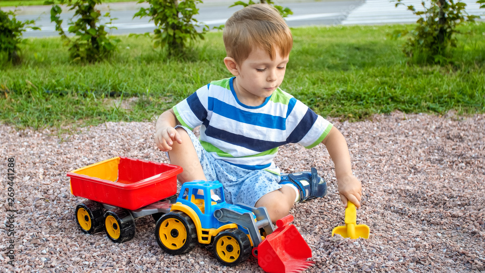Closeup portrait of happy smiling 3 years old child boy digging sand on the playground with toy plastic truck or excavator. Child palying and having fun at park at summer Closeup portrait of happy smiling 3 years old child boy digging sand on the playground with toy plastic truck or excavator. Child palying and having fun at park at summer