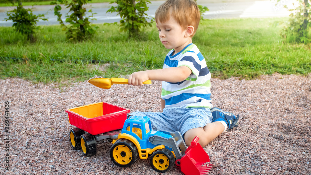 Portrait of adorable 3 years old toddler boy playing with toy truck with trailer on the playground at park. Child digging and building from sand