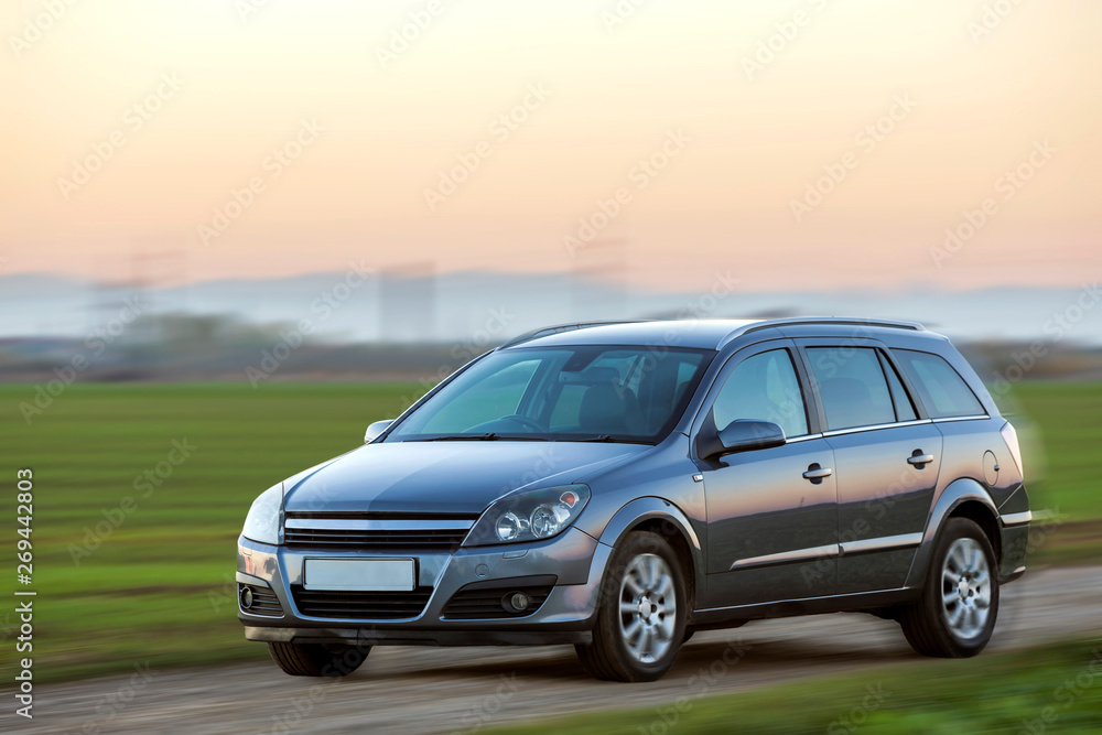 Gray silver new shiny car moving on empty rural road on blurred green meadows and clear foggy sky copy space background. Transportation, speed, comfort and long-distance traveling concept.