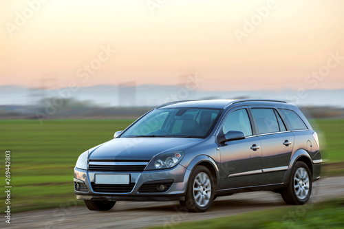 Gray silver new shiny car moving on empty rural road on blurred green meadows and clear foggy sky copy space background. Transportation, speed, comfort and long-distance traveling concept. © bilanol
