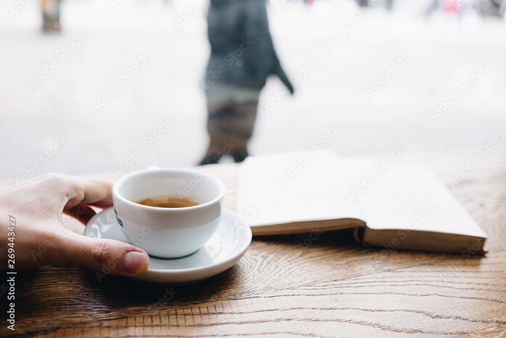 Human hand holding black coffee on saucer with vintage book on background on the wooden table next to big coffee shop window and looking to passerby street walkers Human hand holding black coffee on saucer with vintage book on background on the wooden table next to big coffee shop window and looking to passerby street walkers
