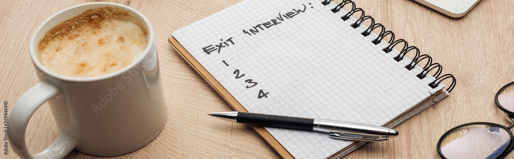 panoramic shot of notebook with exit interview lettering and numbers on wooden table with pen, glasses and cup of coffee panoramic shot of notebook with exit interview lettering and numbers on wooden table with pen, glasses and cup of coffee