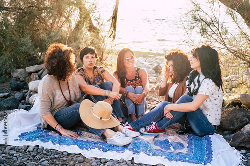 Group of young adult caucasian people women sit down in outdoor leisure activity together at the beach enjoying the sunset light in friendship - friends in beautiful place concept