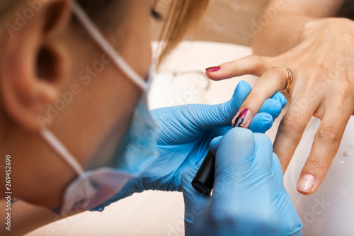 A manicurist in a mask and blue gloves puts a red gel polish on the girl s nails