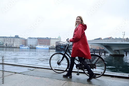 Denmark, Copenhagen, happy woman pushing bicycle at city harbor in rainy weather