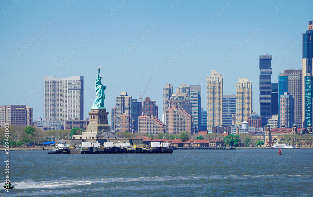 Panorama view of The Statue of Liberty in foreground and New York City skyscrapers in background, Boats in river with blue sky.