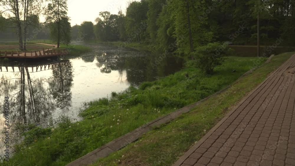 Early morning at the riverbank in a Dobele town, Latvia. Trees foliage reflections in the calm river water. Stone pavement path in a park with a viewing area. Warm sunlight in summer. Early morning at the riverbank in a Dobele town, Latvia. Trees foliage reflections in the calm river water. Stone pavement path in a park with a viewing area. Warm sunlight in summer.