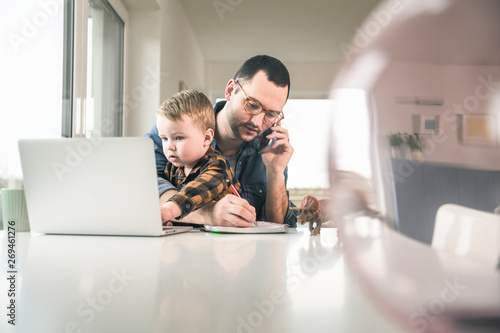 Busy father working at table in home office with son sitting on his lap photo