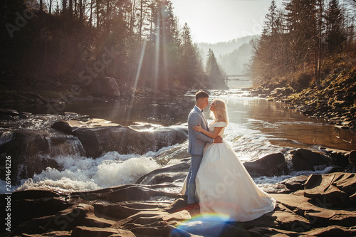 Amazing wedding couple, bride and groom holding hands on a mountains and rivers background. Cute girl in white dress, men in business suit. mountains landscape in the sunset. Concept of family