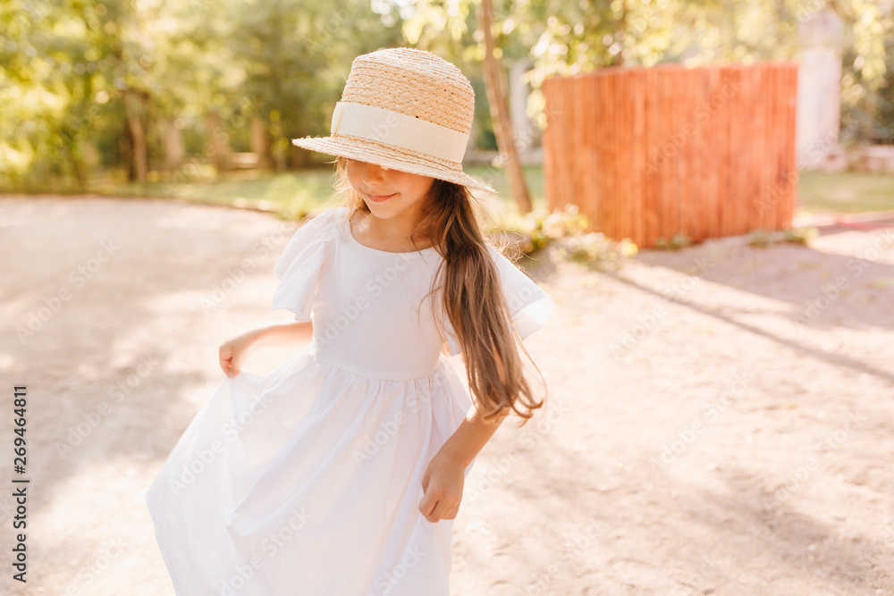 Wonderful little fashionable lady in boater decorated with white ribbon dancing and looking down. Outdoor portrait of tanned girl in long dress spending time in the yard.