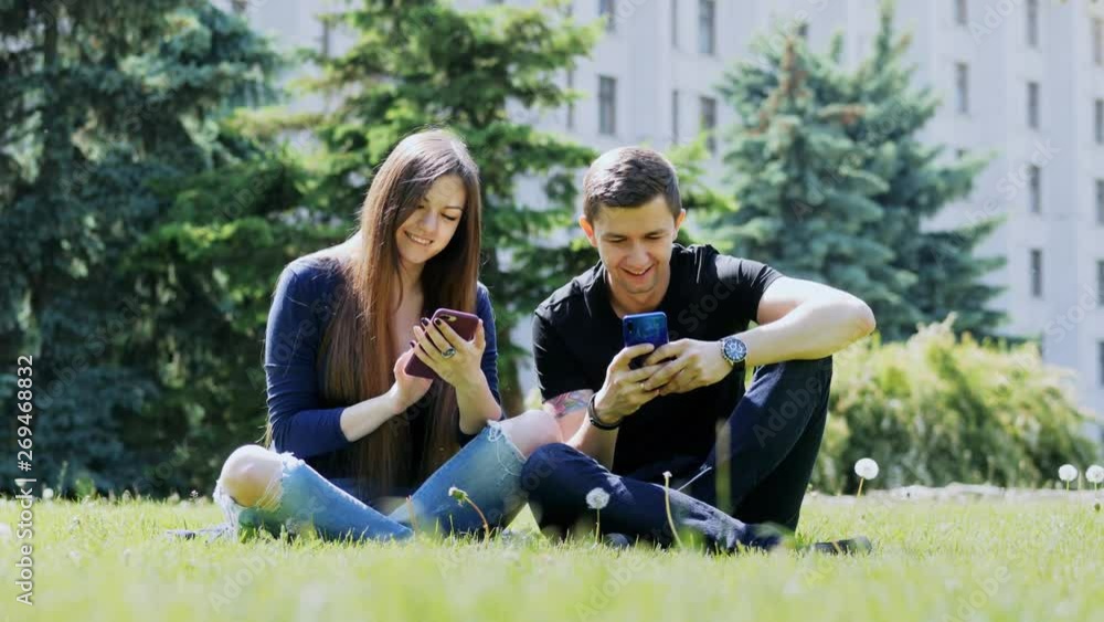 Happy friends use their phones while relaxing in a city park on the grass, chatting, surf iternete Happy friends use their phones while relaxing in a city park on the grass, chatting, surf iternete