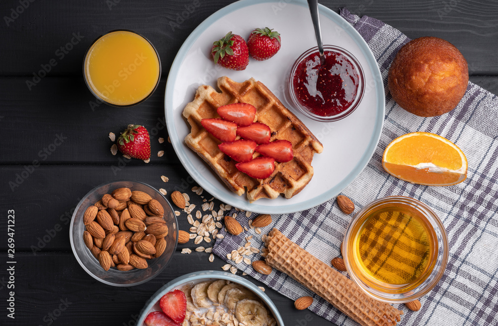 Breakfast. Morning meal set. Delicious oatmeal granola with berries, nuts and honey. A crispy waffle topped with strawberry jam. On a black table background. Top view Breakfast. Morning meal set. Delicious oatmeal granola with berries, nuts and honey. A crispy waffle topped with strawberry jam. On a black table background. Top view