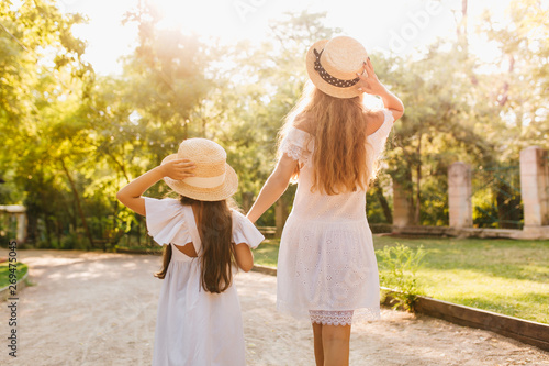 Outdoor portrait from back of tanned woman in white dress and her daughter in similar outfit enjoying lovely nature views. Pretty long-haired lady holding hands with little girl posing in sunlight. © Look!