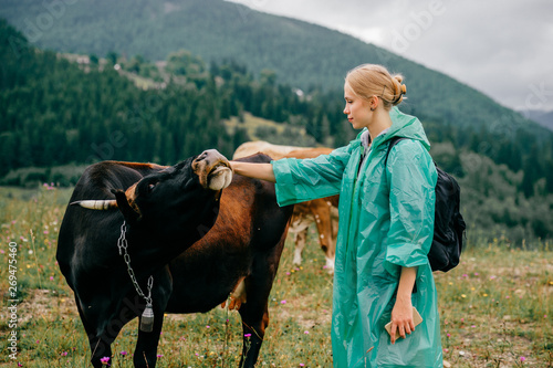 Young beautiful blonde girl in raincoat petting wild bull on pasture in summer cloudy day with landscape view at mountains, hills and forest. Cute woman make friendship with dangerous ox at nature. photo