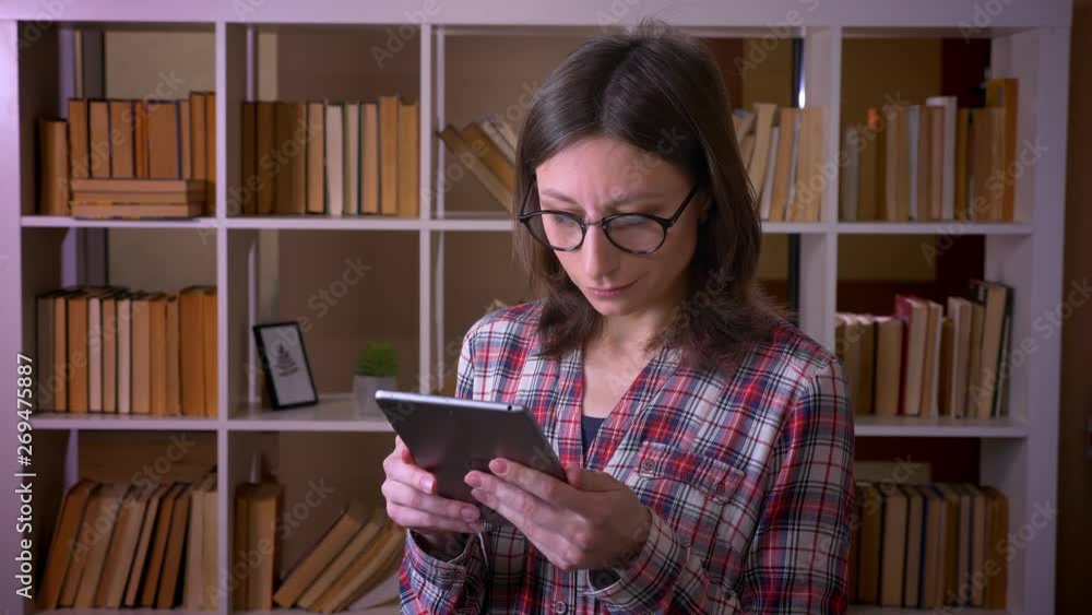 Closeup shoot of young attractive female student in glasses using the tablet and looking at camera smiling in the university library indoors Closeup shoot of young attractive female student in glasses using the tablet and looking at camera smiling in the university library indoors