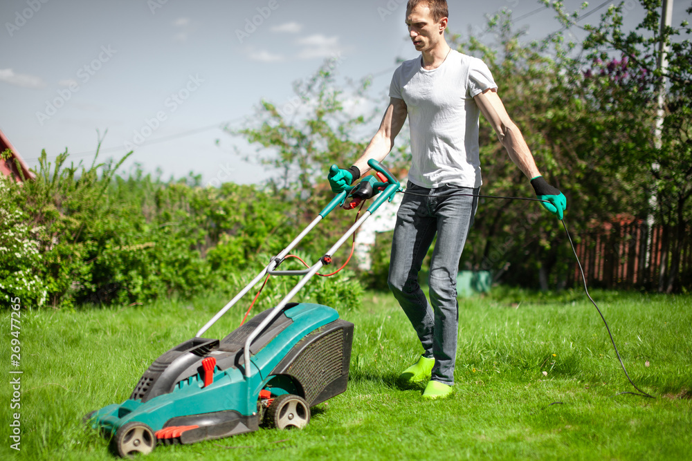 Young man mowing the lawn. Worker doing his job in backyard. Spending summer day in garden. Lawnmower standing on the background of private garden. Young man mowing the lawn. Worker doing his job in backyard. Spending summer day in garden. Lawnmower standing on the background of private garden.