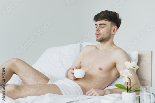pensive handsome man holding man holding coffee cup while lying on white bedding in underwear