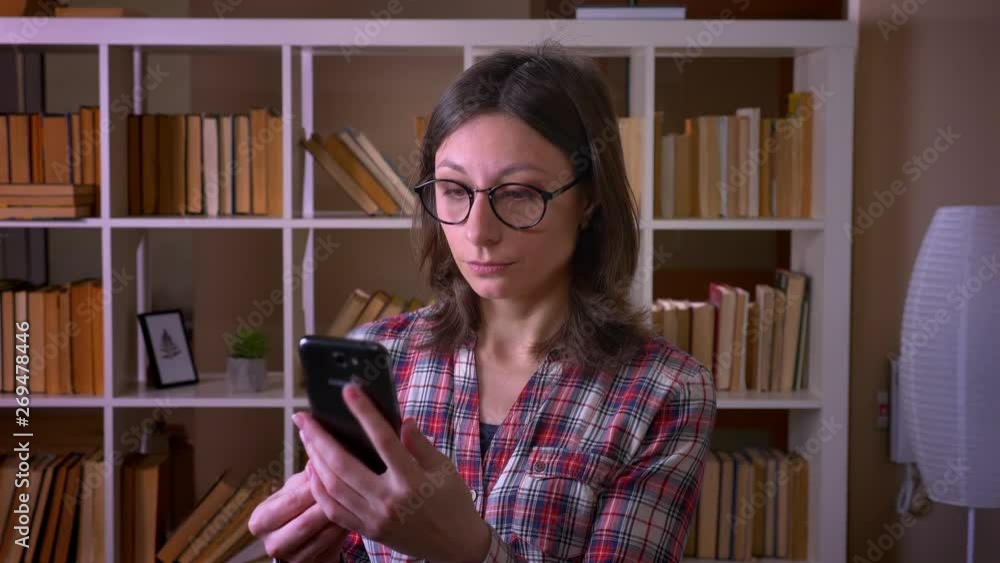 Closeup shoot of young attractive female student in glasses having a video call on the phone in the library indoors Closeup shoot of young attractive female student in glasses having a video call on the phone in the library indoors