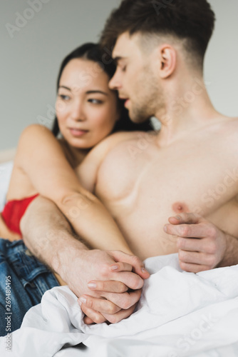 selective focus of loving interracial couple holding hands while lying on bed