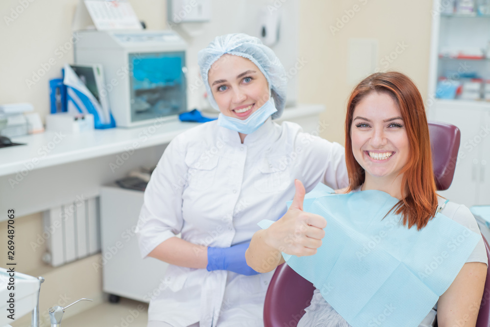 Obraz premium Happy women a dentist and patient after treating teeth at the dental office, smiling and looking towards the camera. Happy patient shows thumb as an endorsement. Dental equipment.